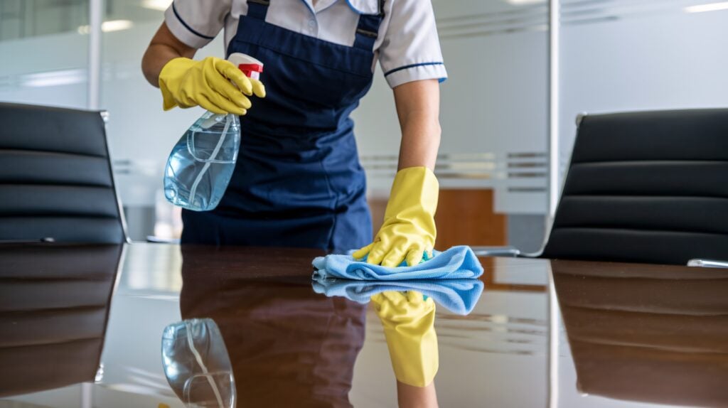 Office Cleaning Staff Sanitizing Conference Table. A dedicated cleaning professional in a blue uniform and yellow gloves meticulously cleans a glass table in a contemporary office setting.
