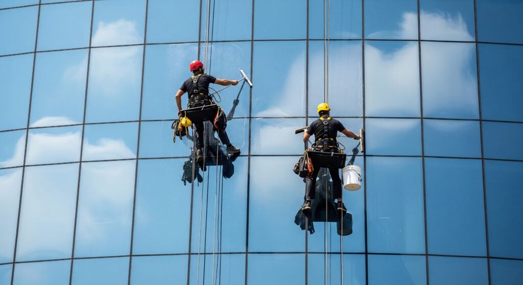 Two professional high-rise window cleaners suspended by ropes, meticulously cleaning the reflective glass facade of a modern office building on a clear sunny day.